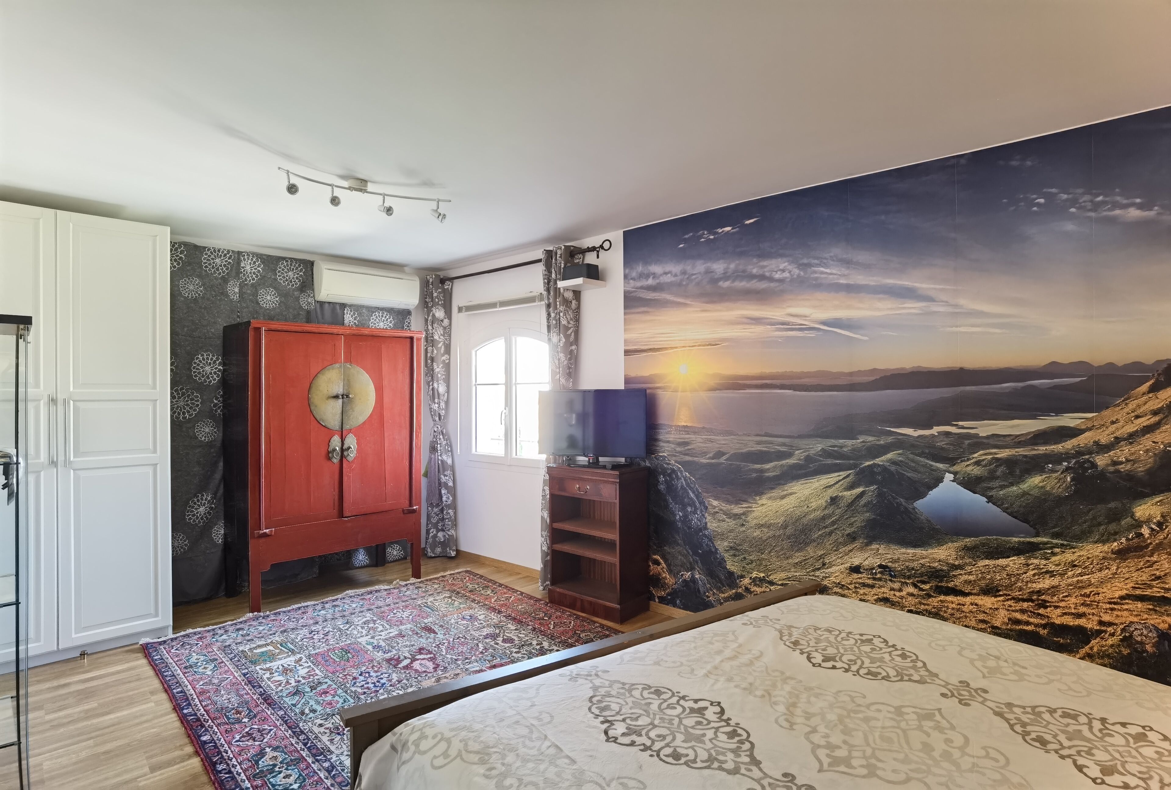 A bedroom with a large bed, a red wardrobe, a television on a wooden stand, and a scenic sunset mural on the wall.