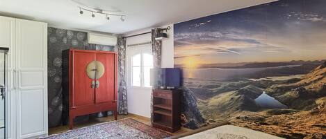 Un dormitorio con una cama grande, un armario rojo, un televisor sobre un soporte de madera y un mural con atardecer panorámico en la pared.