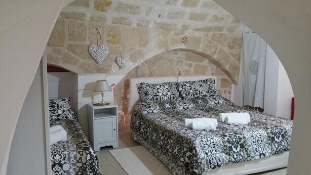 A bedroom with a stone wall, a bed with a patterned bedspread, a nightstand with a lamp, and heart-shaped decorations. - Quad room (Monopoli)