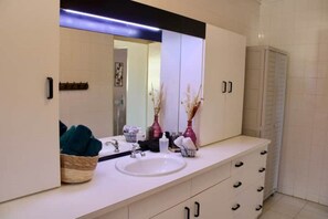 Une salle de bain moderne avec un grand miroir, un meuble-lavabo blanc et un évier. On y trouve des vases décoratifs sur le meuble-lavabo et un panier rempli de serviettes.