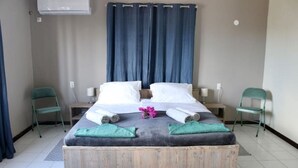 Une chambre d’hôtel avec un lit en bois, deux chaises vertes, une petite table et un rideau bleu.