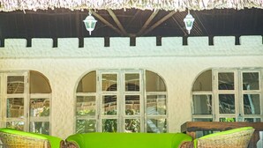 Un espace extérieur aménagé, abrité et doté d’un canapé vert et de fauteuils en osier, de grandes fenêtres en arc de cercle et de lanternes suspendues.