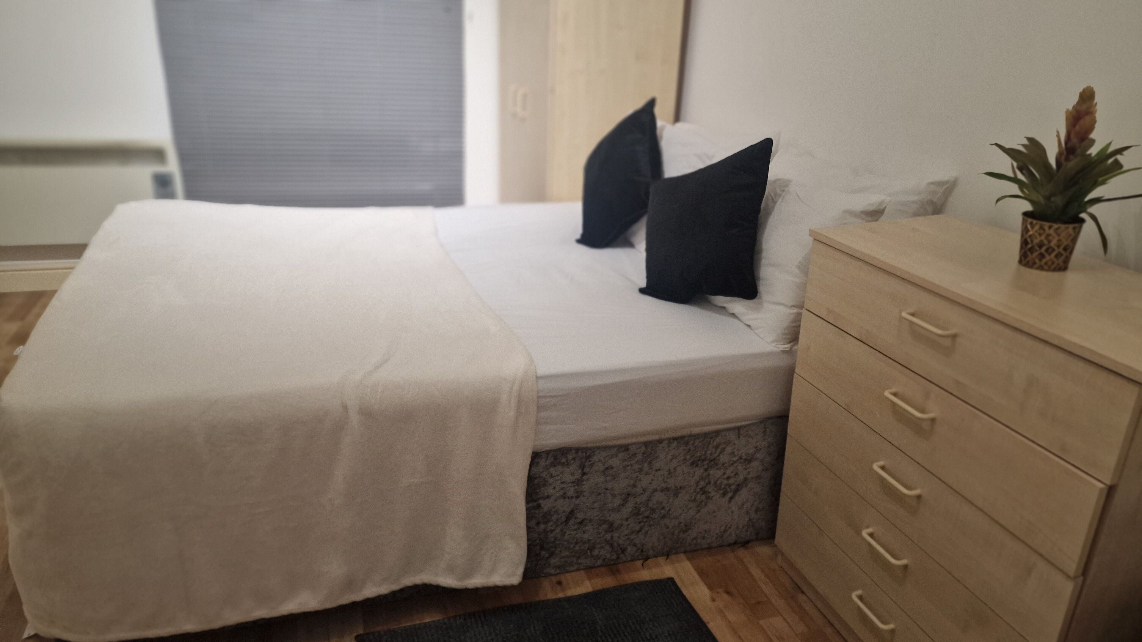 Ein Schlafzimmer mit Bett, Kommode, Pflanze und Fenster mit Jalousien.