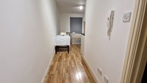 A narrow hallway with wooden flooring, a small table with a white tablecloth, a bed with a grey headboard, and a wall-mounted telephone. - Compact Living Cosy Studio Flat in London (London)