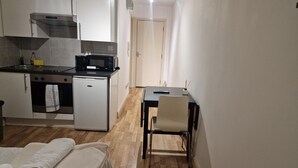 A compact kitchen with a refrigerator, microwave, and stove. There's a small dining table with a chair and a remote control on it. A towel hangs on the oven door. - Compact Living Cosy Studio Flat in London (London)