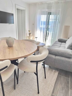 A modern living room with a round wooden dining table, beige upholstered chairs, a grey sofa, and a TV mounted on the wall. - Holiday flat with terrace and sea view (Zadar - Kruševo Karin)