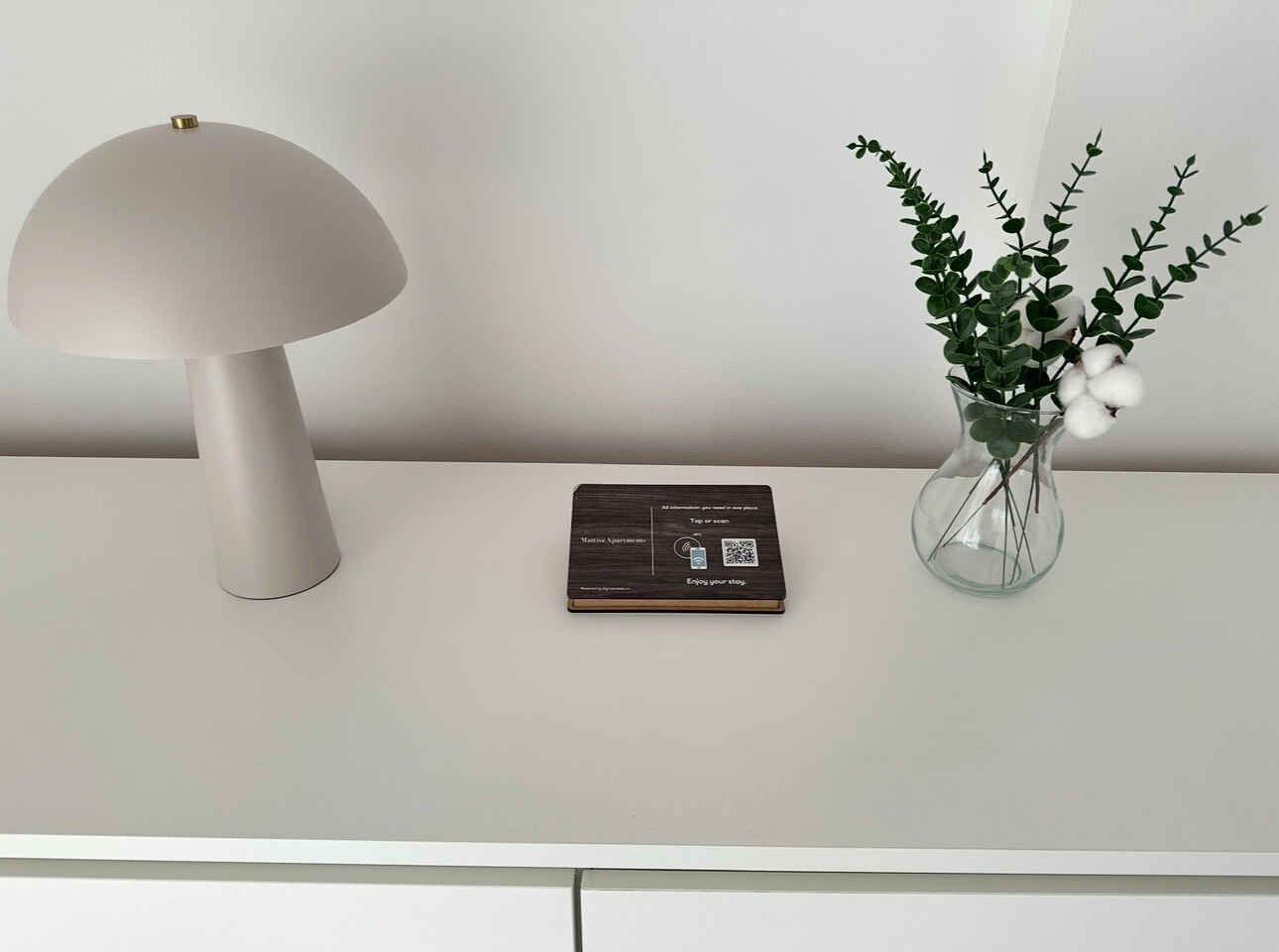 Una mesa moderna con una lámpara blanca, una placa con un código QR y un florero con plantas verdes y flores blancas.