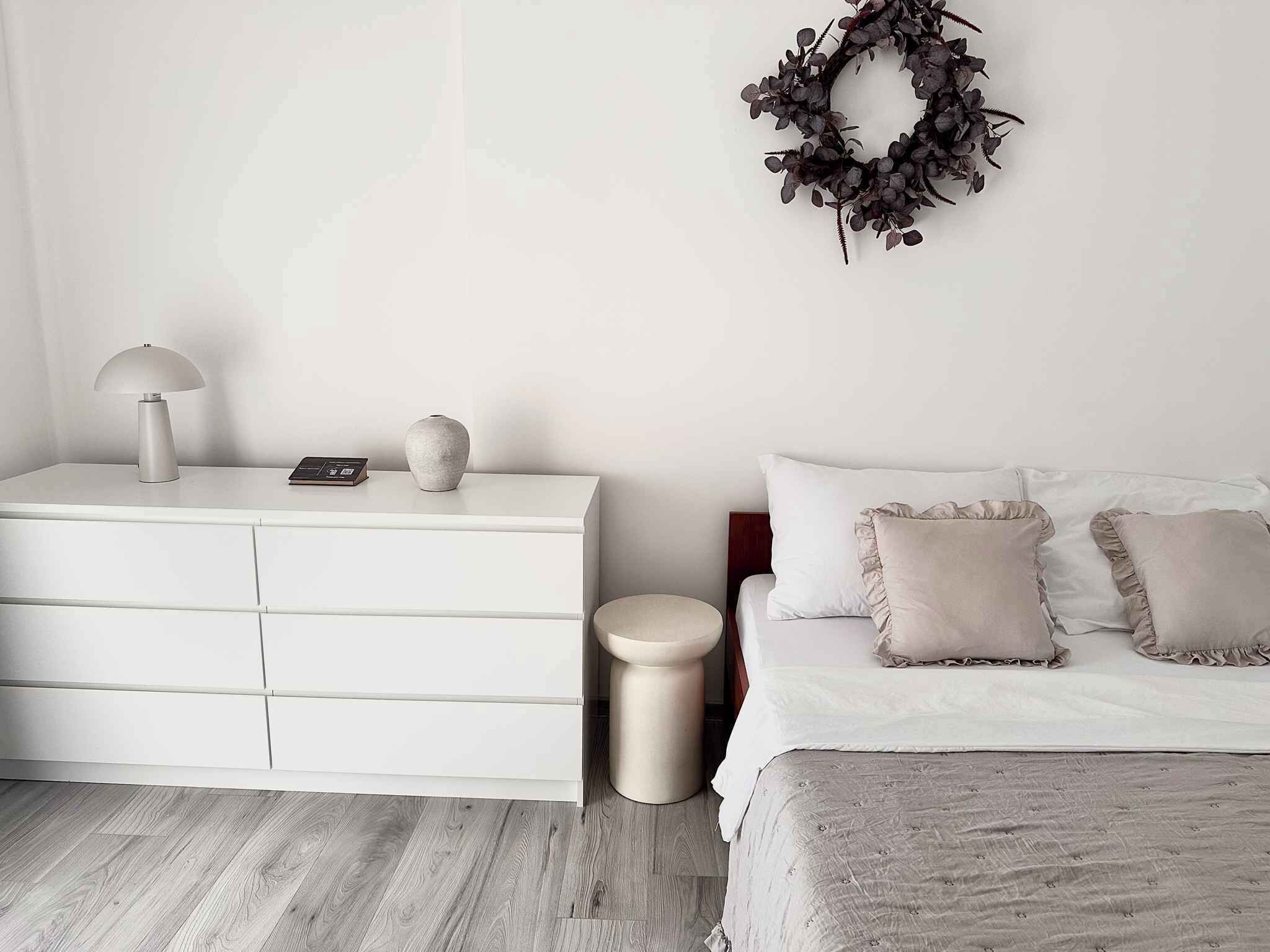 Un dormitorio minimalista con un tocador blanco, una mesita de noche, una lámpara, un jarrón pequeño, un taburete redondo, una cama con almohadas con volantes y una guirnalda oscura en la pared.