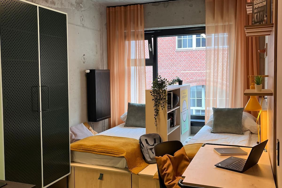 A compact, well-lit room with a bed, a desk with a laptop, a chair, a small rug, and a window with curtains.