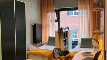 A compact, well-lit room with a bed, a desk with a laptop, a chair, a small rug, and a window with curtains.