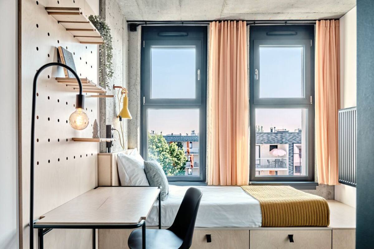 A compact room with a bed, a desk, and a chair. There are two windows with curtains, a wall-mounted shelf with a lamp, and a radiator.