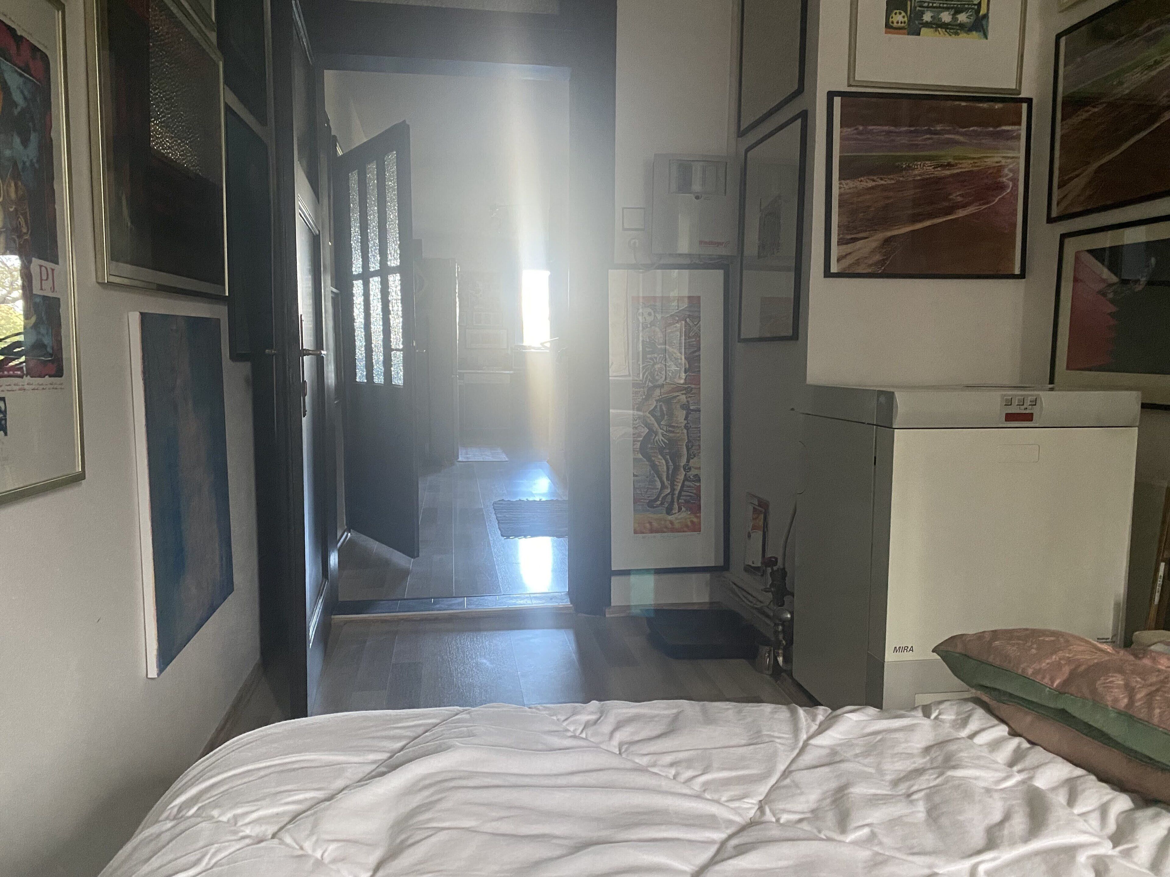 Um corredor estreito que leva a um quarto com uma cama, um refrigerador e várias quadros nas paredes.