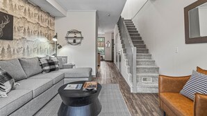 Una sala de estar moderna con un sofá gris, una mesa de centro negra y un sillón de cuero marrón. Una escalera con barandilla de madera lleva a un piso superior.