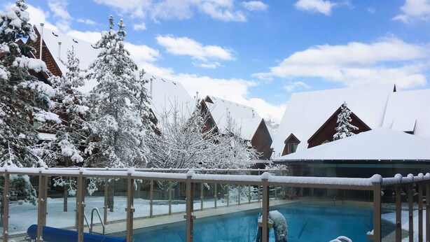 Una piscina exterior cubierta de nieve, con sillas de descanso y una barandilla de vidrio.