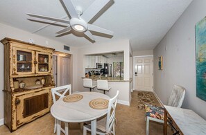 Un comedor con una mesa y sillas blancas, un ventilador de techo y una cocina al fondo.