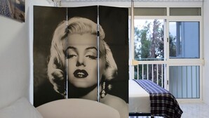 A room with a bed, a window with a view of trees, a balcony with a railing, and a large poster on the wall.