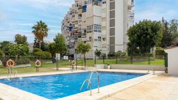 A swimming pool with a metal ladder, surrounded by a fence with lifebuoys, and a tall residential building in the background.
