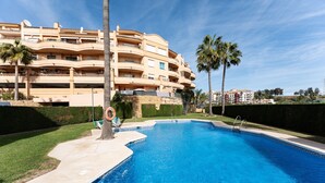 A swimming pool with a palm tree, a lifeguard chair, and a building with balconies in the background. - Holiday Apartment "Penthouse 3-Bedroom Duplex" with Sea View, Shared Pool & Wi-Fi (Mijas)