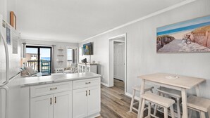 A kitchen with white cabinets, a dining area with a table and benches, and a painting of a beach scene on the wall. - Ocean Forest Villas Unit B305 - Ocean view (Myrtle Beach)