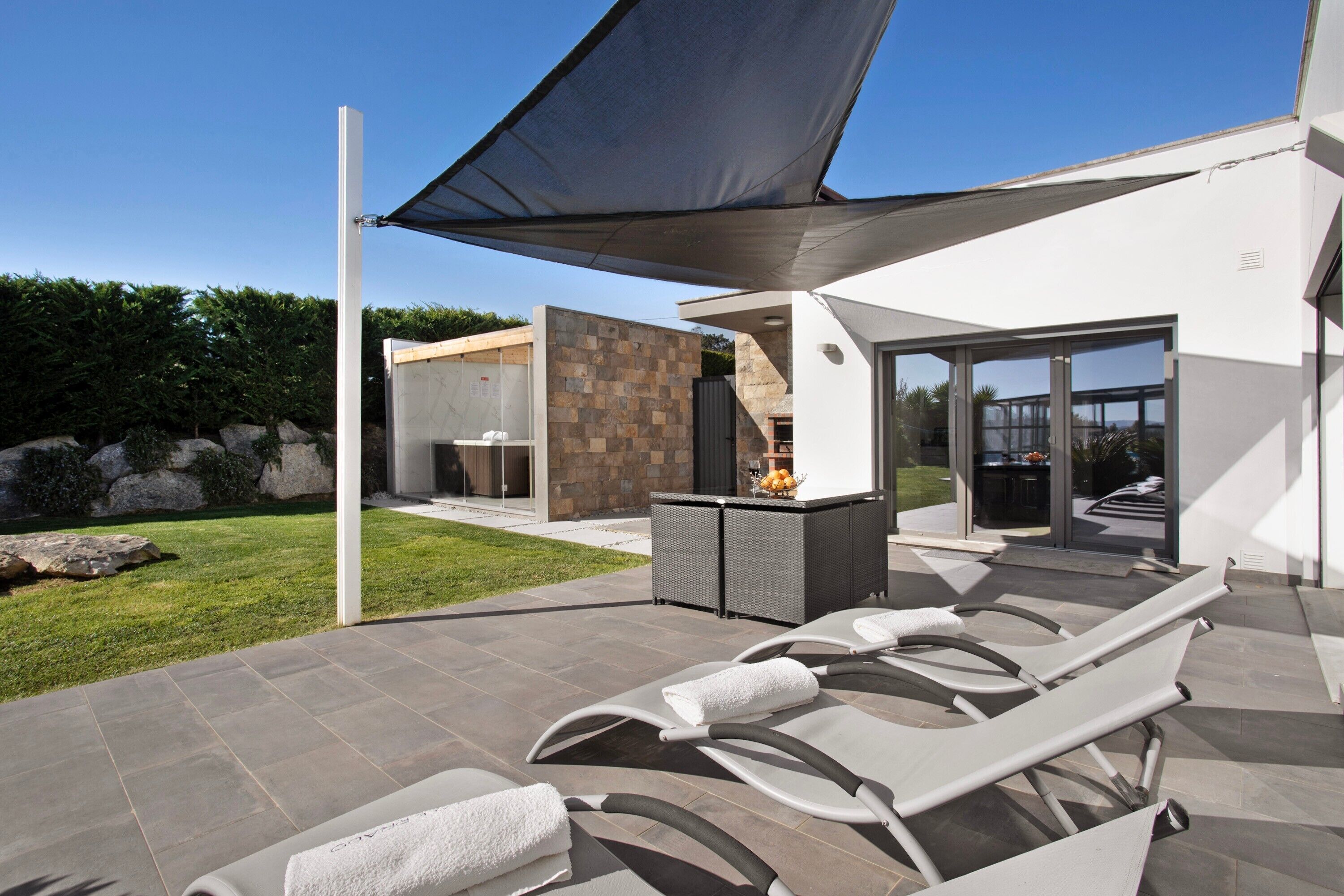 Un área de lounge exterior moderna con tumbonas, un muro de piedra y una puerta de cristal que da a un patio.