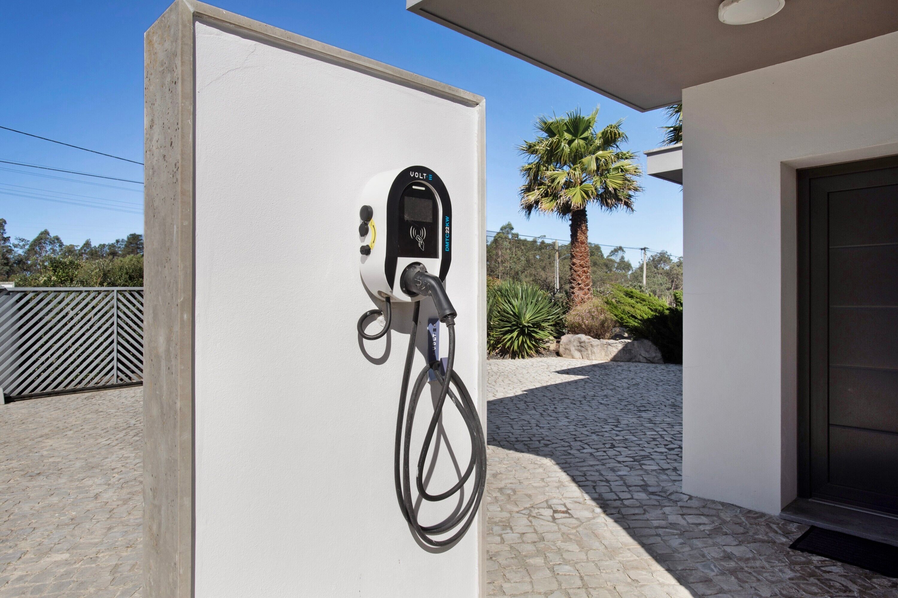 An electric vehicle charging station attached to a white wall, with a palm tree and a modern building in the background.