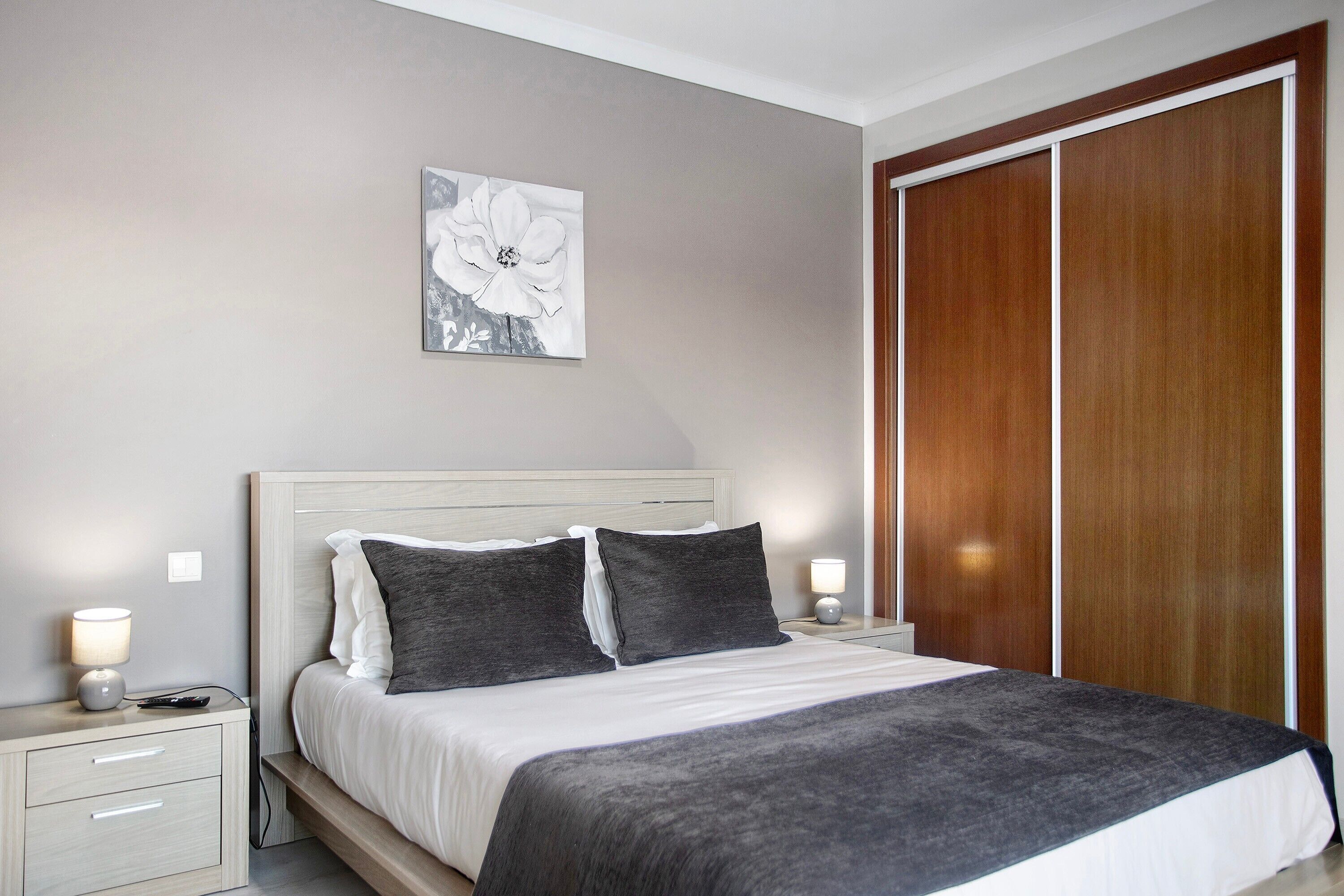 A bedroom with a bed, bedside tables, a nightstand, a wall-mounted lamp, and a wooden wardrobe.