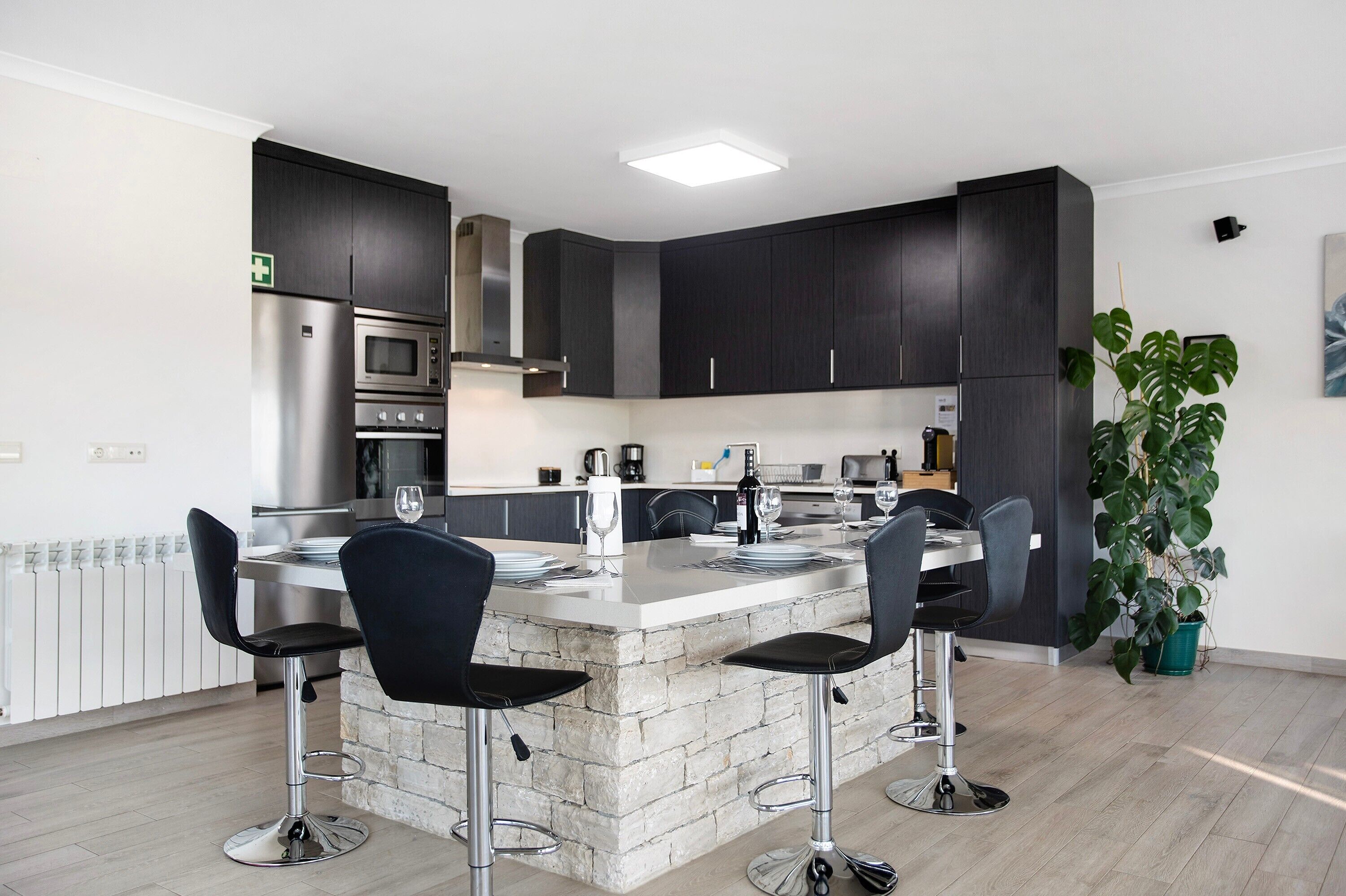 Una cocina moderna con armarios oscuros, una isla central con taburetes de bar, electrodomésticos de acero inoxidable y una planta en maceta.