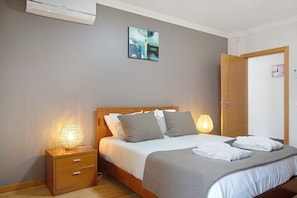 Une chambre à coucher comprenant un lit, des lampes de chevet, une table de chevet, une tête de lit en bois, un climatiseur mural et une porte.