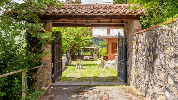 An entrance with a tiled roof, a stone wall, and a gate leading to a courtyard with potted plants and a building in the background. - Holiday Home 'Seaview Camaiore' with Sea View, Private Pool and Wi-Fi (Camaiore)