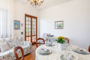 A dining area with a table set for four, chairs, a sofa, a chandelier, and a door with glass panels.