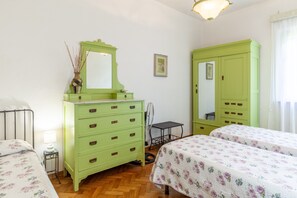 A room with two beds, a green dresser with drawers, a mirror, a small table, and a fan.