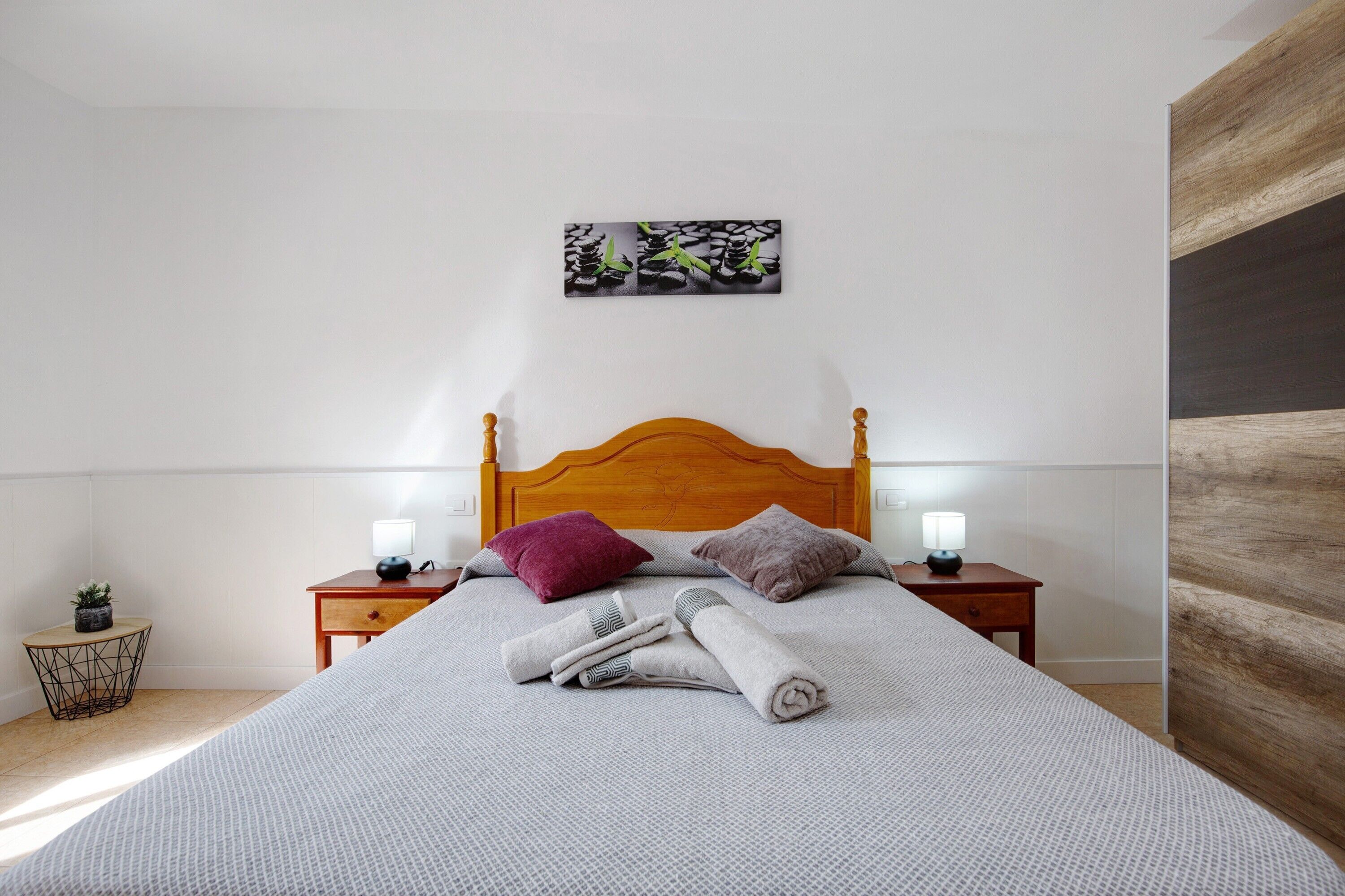 Una cama bien hecha con almohadas, una mesita de noche con lámpara, una pequeña mesita con una planta y una obra de arte en la pared.