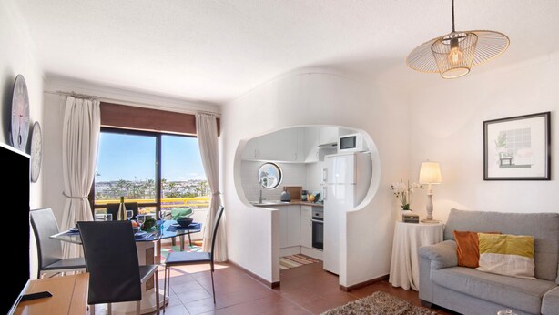 A modern living room with a sofa, a dining table with chairs, a kitchen area, and a view through a large window. - Apartment 'Montechoro' with Balcony and Wi-Fi (Albufeira)