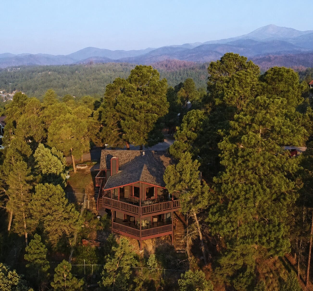 A multi-level wooden cabin surrounded by dense forest with a mountain range in the background.