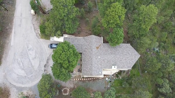 An aerial view of a house with a sloped roof, surrounded by dense greenery and a dirt road.
