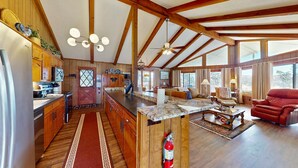 A spacious kitchen with wooden cabinets, a marble countertop, and a large island. There is a dining area with a red sofa and a coffee table. The room has wooden beams on the ceiling and large windows.