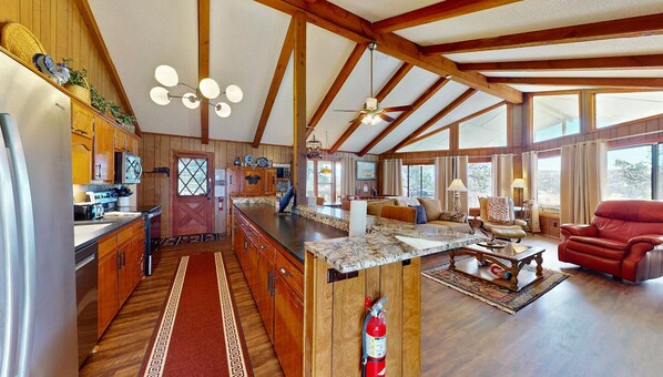 Una cocina amplia con armarios de madera, encimera de mármol y una isla grande. Hay una zona de comedor con un sofá rojo y una mesa de centro. La habitación tiene vigas de madera en el techo y ventanas amplias.