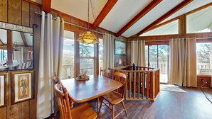 A dining area with a wooden table and chairs, a hanging light fixture, and large windows offering a view of the outdoors.