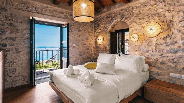 A bedroom with a stone wall, a bed with white linens, a wooden ceiling, and a balcony with a view. - Pyrgos Towers (West Mani)