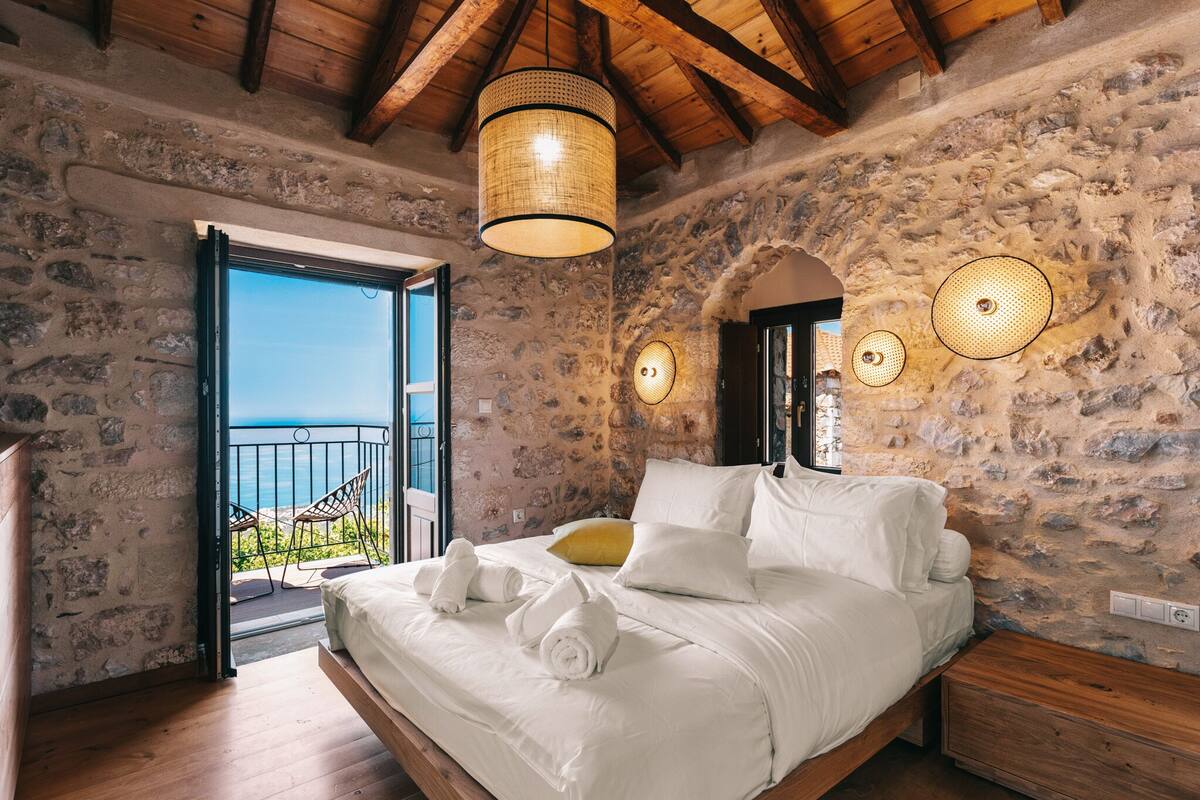 A bedroom with a stone wall, a bed with white linens, a wooden ceiling, and a balcony with a view.