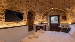 A bedroom with a stone wall, a flat-screen TV mounted on the wall, a bed with towels, a small table, and a window with blinds. - Pyrgos Towers (West Mani)