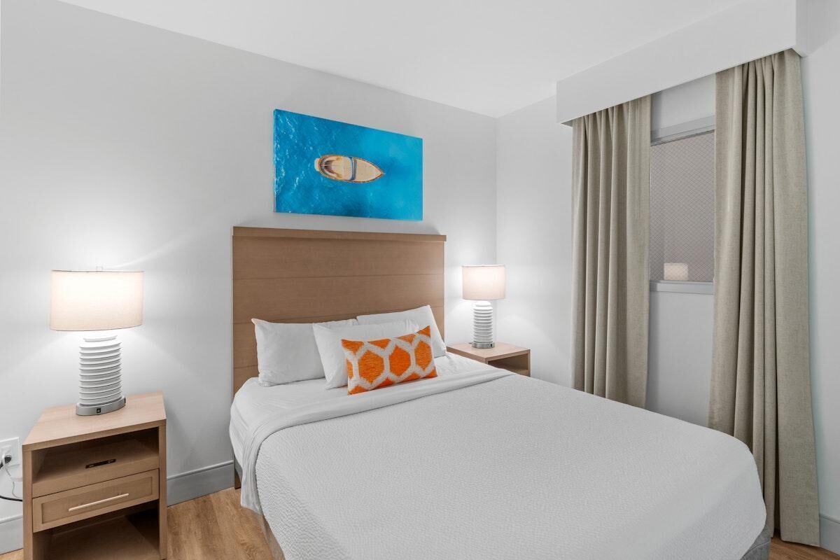Une chambre d’hôtel avec un grand lit, deux tables de chevet avec des lampes, une tête de lit, une fenêtre avec des rideaux et une œuvre d’art bleue au-dessus du lit.
