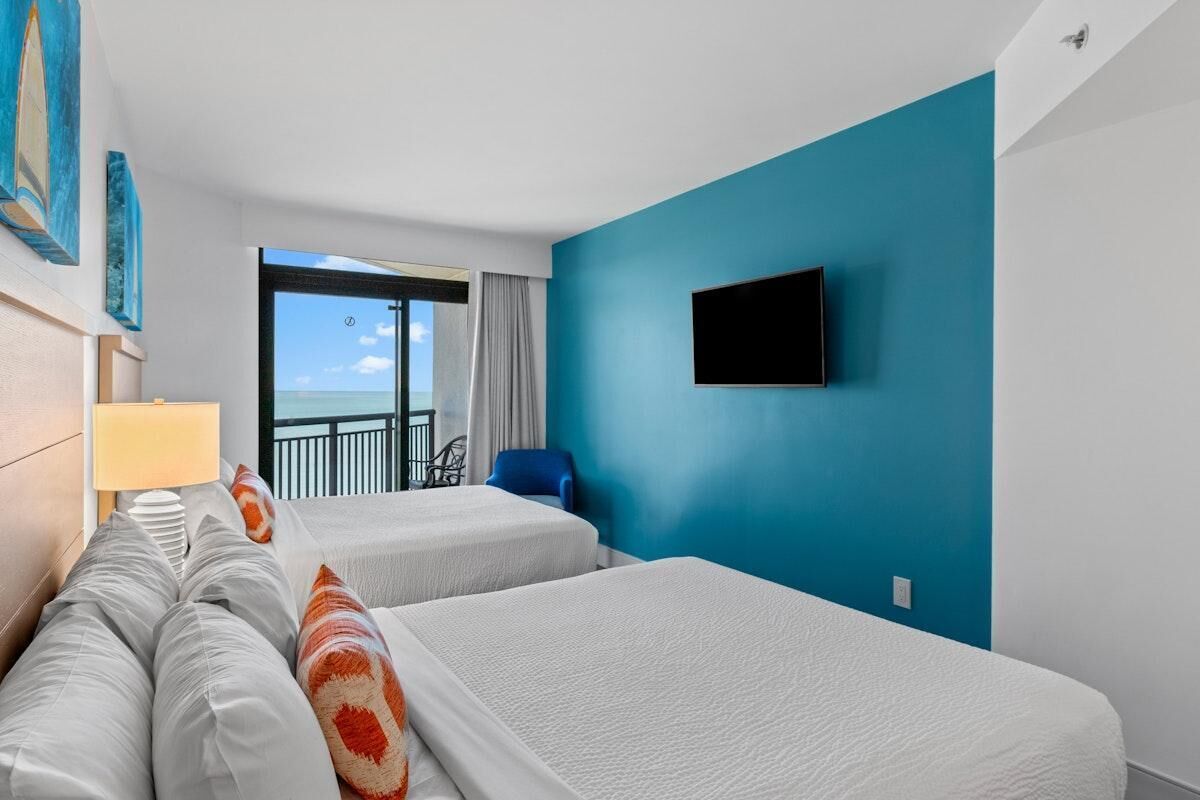 Habitación de hotel con dos camas, televisión, balcón con vistas y una silla.