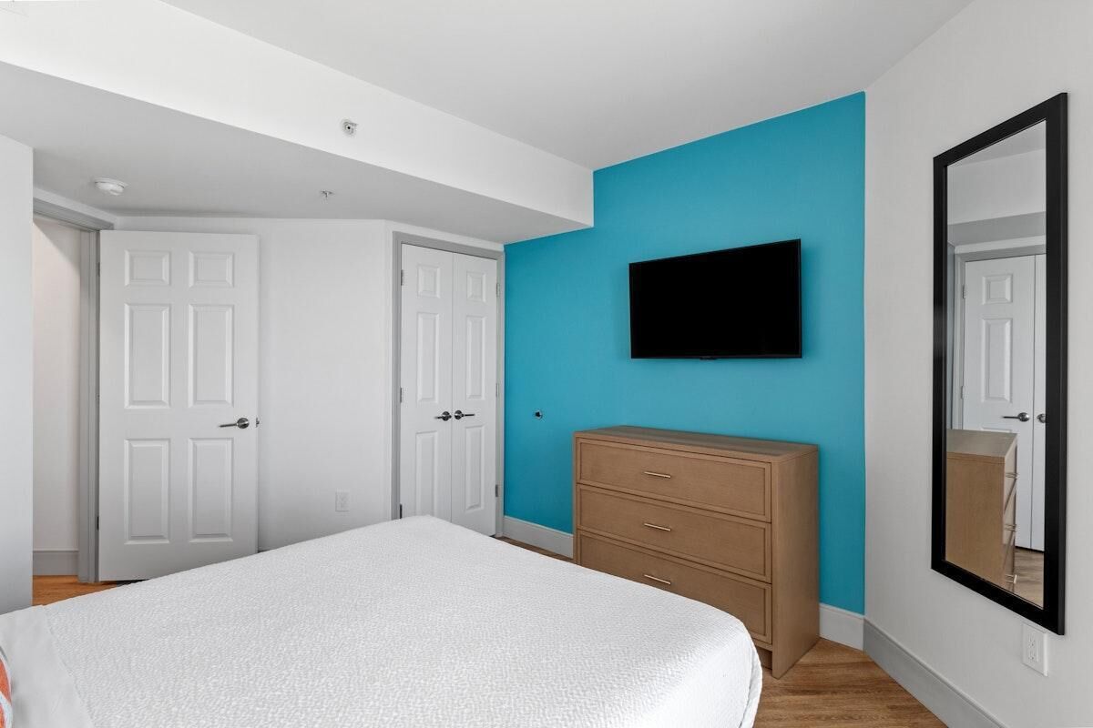 Un dormitorio con cama, cómoda, televisor en la pared y dos puertas blancas.