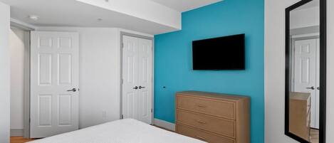 Un dormitorio con cama, cómoda, televisor en la pared y dos puertas blancas.