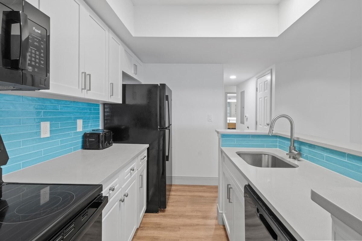 Een moderne keuken met witte kastjes, een zwarte koelkast, een magnetron en een blauwe tegelachtergrond.