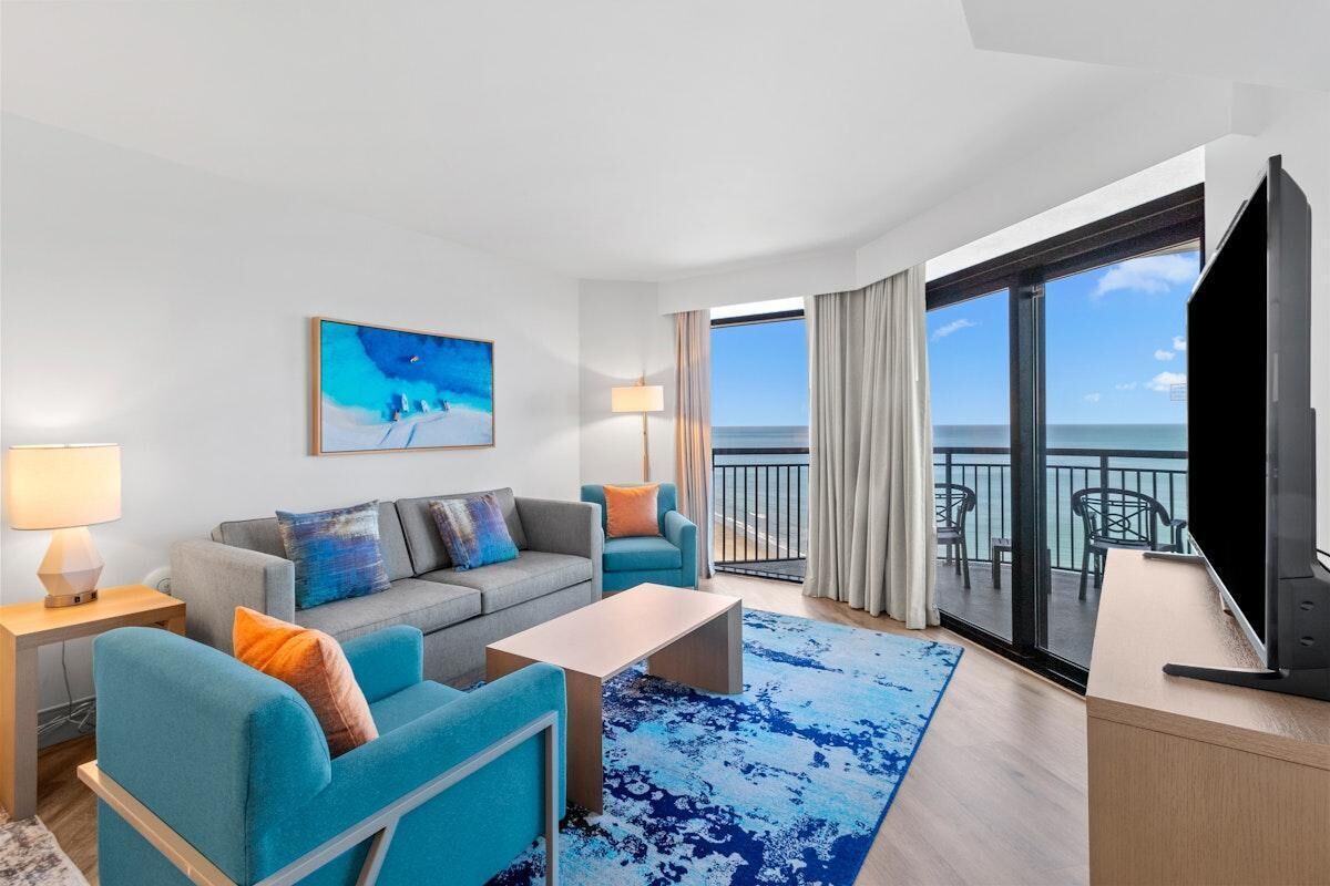 A modern hotel room with a balcony, a sofa set, a coffee table, and a TV.