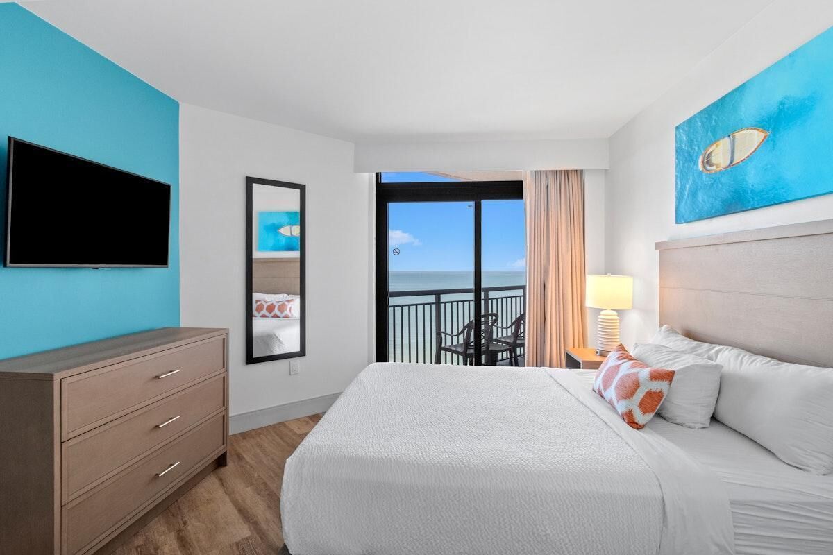 A hotel room with a large bed, a flat-screen TV, a dresser, and a balcony with a view of the ocean.