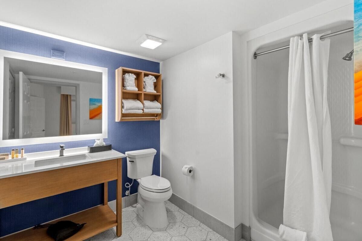 Un baño con un lavamanos blanco, un inodoro, una ducha con cortina blanca y un estante con toallas dobladas.