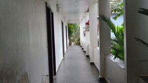 A long, narrow corridor with white walls and a dark floor, leading to a covered area with plants and a red structure. - Psygroover Rococco (Mandrem)